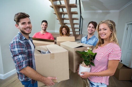 A group of five young adults, three men and two women, gathered inside a bright, modern house with a neutral-toned wall and a wooden staircase in the background, preparing for a house move. The individuals are smiling and holding various household items and packed cardboard boxes, indicating they are in the process of moving. One woman in a pink striped shirt is holding a small potted plant with green leaves, while another man wearing a checked shirt supports a large cardboard box filled with household belongings. The scene suggests a friendly and organized home removal in Hammersmith or nearby West London, with the group standing around a spacious interior with natural light and a tidy, minimalist style, reflecting the typical environment for local removals and house relocation services offered by Hammersmith Man with Van.