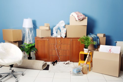 The image depicts a room with a light blue wall and a white tiled floor, cluttered with various household items and packing supplies. In the center, there is a wooden sideboard or cabinet with a reddish-brown finish, topped with several cardboard moving boxes in different sizes, some sealed with packing tape and others open, revealing household items inside. A clear plastic bag containing wrapping material is leaning against the sideboard, while a red and white checkered cloth is draped over one of the boxes. To the right of the sideboard, a small yellow shelving unit holds a potted green plant, and a cardboard box sits nearby, partially open with packing paper visible. A rolled-up beige rug lies on the floor in front of the sideboard, along with some loose packing materials and a pair of black shoes. Scattered across the scene are additional cardboard boxes, some partially filled, and various household items like a plastic container. The space appears to be a living room or a hallway in the midst of packing for a move, with natural daylight illuminating the cluttered environment and reinforcing the context of house removals in Hammersmith or nearby areas.