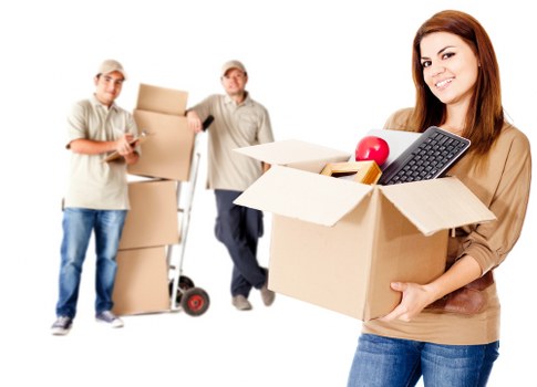 A young woman with long brown hair, dressed in a light brown top and blue jeans, holding an open cardboard moving box filled with household items such as a red apple, a black remote control, a yellow sponge, and a small stack of papers, smiling at the camera. In the background, two men, both wearing beige work shirts and casual jeans, are standing in a residential setting with their own boxes and equipment: one man is holding a box and the other is pushing a small trolley loaded with cardboard boxes. The scene suggests a professional house removal or moving service, with the environment appearing to be inside or near a home, indicating a typical house move transition in the Hammersmith area. The lighting is bright and even, emphasizing the organized and systematic nature of the move as provided by Hammersmith Man with Van, a local removals specialist.