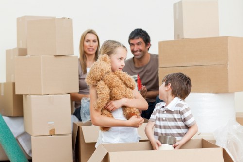 A family of four, including a young girl holding a fluffy tan teddy bear and a boy wearing a striped shirt, stand inside a bright, spacious room surrounded by numerous cardboard boxes of various sizes, some closed and others open, revealing household items. The boxes are plain brown with a textured cardboard finish, and are stacked in the background and foreground, indicating a moving or packing process. The family members appear relaxed and engaged, with the adults smiling and observing their children. The scene is well-lit with natural light, and the setting looks like a residential interior, possibly an entrance hall or living room, in Hammersmith or nearby in West London. The presence of the boxes and a packed, utilitarian environment aligns with house removals and relocation services, with Hammersmith Man with Van's branding subtly implied to match the local context of the postcode area.