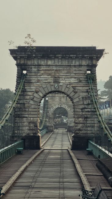 A historic stone arch bridge with a roadway and railway tracks running through its center, supported by green metal suspension cables attached to sturdy stone pillars. The bridge, partially obscured b