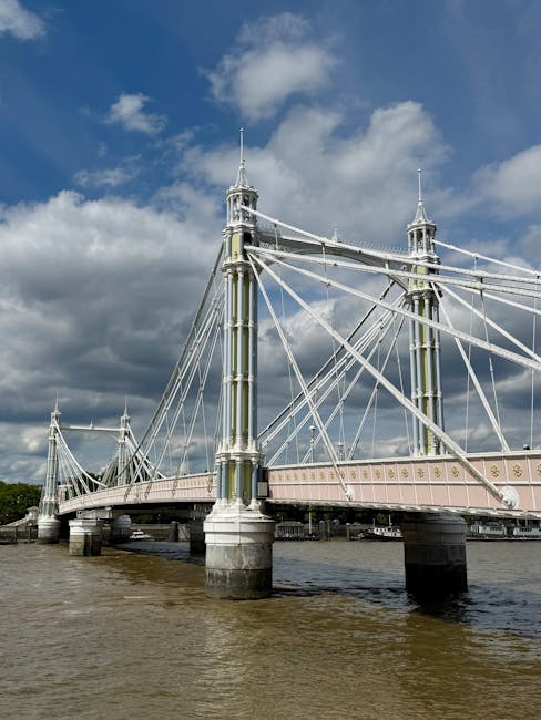 A historic stone arch bridge with a roadway and railway tracks running through its center, supported by green metal suspension cables attached to sturdy stone pillars. The bridge, partially obscured b