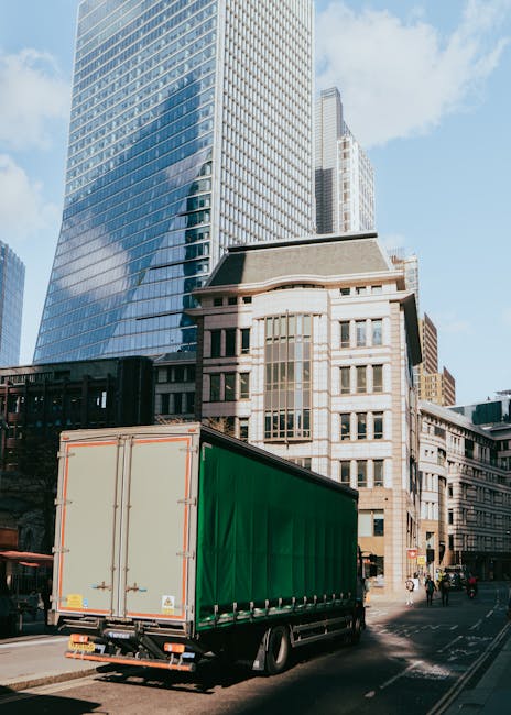 A large green moving truck with a closed cargo area is parked on a city street in Hammersmith, London, surrounded by tall modern glass skyscrapers and older stone buildings. The truck sits near the ke